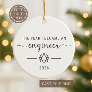 May include: A white ceramic ornament with the text "THE YEAR I BECAME AN engineer" in black script. Below the text is a gear graphic and the year "2026". The ornament hangs from a gold string. A "FREE GIFT BOX" sticker is in the upper left.