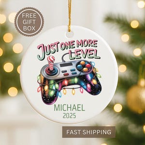 May include: A white ceramic Christmas ornament featuring a colorful video game controller design with the text "Just One More Level". The ornament includes the name "Michael" and the year "2025". A string is attached for hanging.