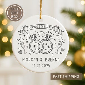 May include: White ceramic ornament with a gold string, featuring a black and white illustration of two diamond rings, champagne glasses, and hearts. Text reads "FOREVER STARTS HERE," and names "MORGAN & BRENNA" with the date "11.21.2025."