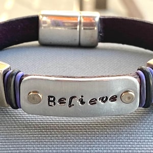 May include: A dark purple leather bracelet featuring a central silver-toned metal plate engraved with the word Believe. The bracelet includes two square silver-toned metal charms depicting horses, accented by small black and purple rings.