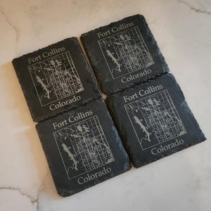 May include: Four square, dark gray slate coasters. Each coaster features a map of Fort Collins, Colorado, with the city name and state name etched in white. The coasters are arranged on a light-colored surface.