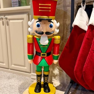 Puede incluir: Una gran figura de cascanueces de colores con uniforme tradicional. La figura presenta un sombrero y una chaqueta rojos, pantalones verdes y botas negras. Se apoya sobre una base circular amarilla, con una cara y un bigote detallados.