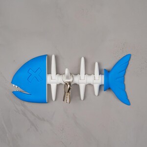 May include: A novelty key holder in the shape of a fish skeleton. The key holder is blue and white, with a cartoon design. It is mounted on a grey wall and holds a set of keys. The fish has a blue head and tail.