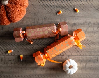 May include: Overhead shot of Halloween-themed party crackers in orange and pink with gold accents, tied with orange ribbons. Candy corn and decorative pumpkins are scattered on a wooden surface.
