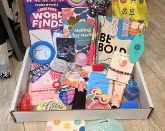May include: A cardboard gift box filled with assorted items including a word search book, a notebook with the text BE BOLD, a sequined star, a keychain, a peppermint face mask, a lip balm, a blue silicone mat, and a card that says You can do HARD THINGS.