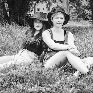 May include: Two people wearing wide-brimmed hats sit in a grassy field. The person on the left is wearing a black shirt and denim shorts. The person on the right is wearing a black tank top and denim overalls.