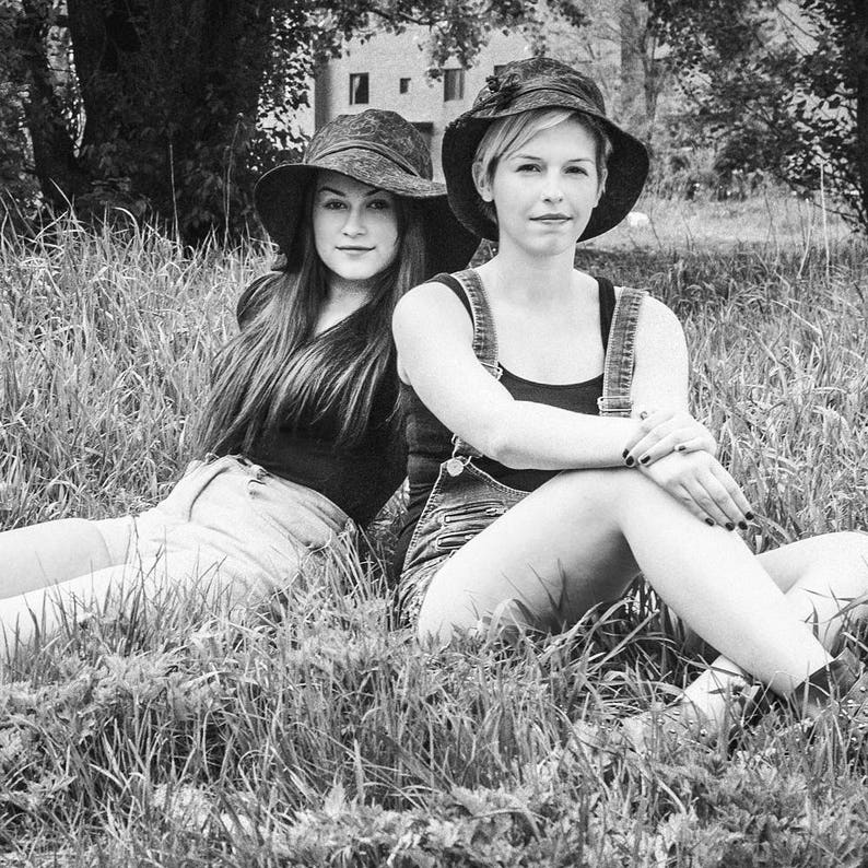 May include: Two people wearing wide-brimmed hats sit in a grassy field. The person on the left is wearing a black shirt and denim shorts. The person on the right is wearing a black tank top and denim overalls.