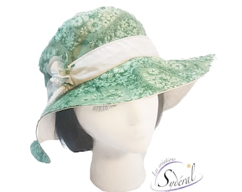 Puede incluir: Un sombrero de sol floral verde con una cinta blanca y un lazo blanco. El sombrero está en una cabeza de maniquí blanca.