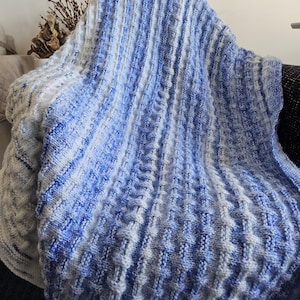 May include: A knitted blanket in shades of blue and white, draped over a chair. The blanket features a textured pattern with vertical and square designs. The color transitions from white to blue, creating a gradient effect. The blanket appears soft and cozy.