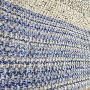 May include: A knitted blanket in shades of white and blue. The blanket has a textured, waffle-like pattern. The color gradient shifts from a darker blue at the bottom to white at the top. The blanket is folded.