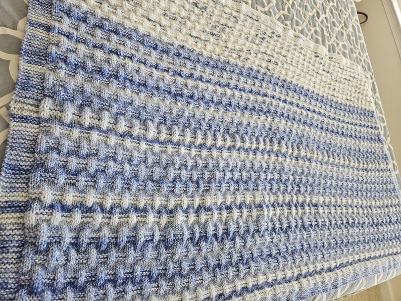 May include: A knitted blanket in shades of white and blue. The blanket has a textured, waffle-like pattern. The color gradient shifts from a darker blue at the bottom to white at the top. The blanket is folded.