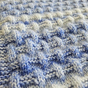 May include: Close-up of a knitted blanket with a textured pattern. The blanket features a combination of white and blue yarns, creating a soft, mottled appearance. The knit stitch creates a raised, dimensional effect, adding visual interest to the textile.