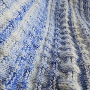 May include: Close-up of a knitted textile with a textured pattern. The fabric features a blend of blue and white yarns, creating a gradient effect. The knitted design has vertical ridges and diamond-shaped details, giving it a three-dimensional appearance.