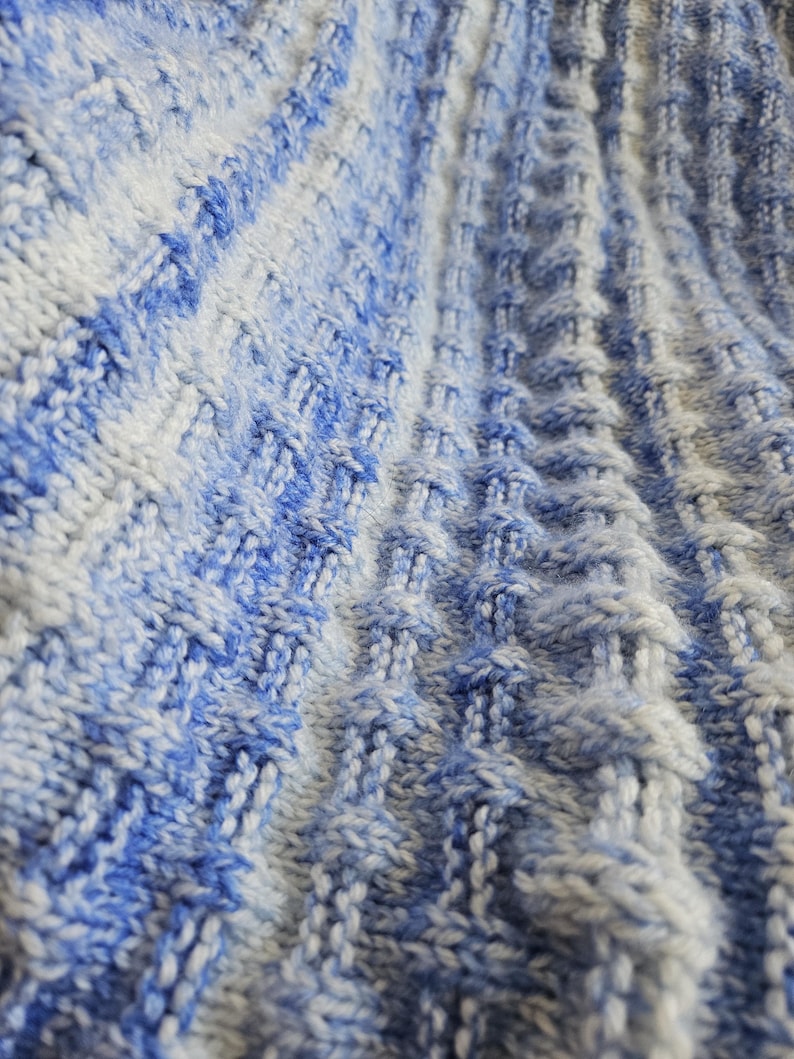 May include: Close-up of a knitted textile with a textured pattern. The fabric features a blend of blue and white yarns, creating a gradient effect. The knitted design has vertical ridges and diamond-shaped details, giving it a three-dimensional appearance.