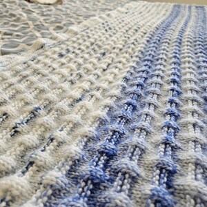 May include: A knitted blanket with a textured pattern in shades of white and blue. The blanket has a gradient effect, transitioning from white to blue. The knitted design creates a raised, dimensional surface.