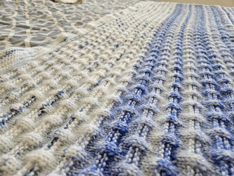 May include: A knitted blanket with a textured pattern in shades of white and blue. The blanket has a gradient effect, transitioning from white to blue. The knitted design creates a raised, dimensional surface.