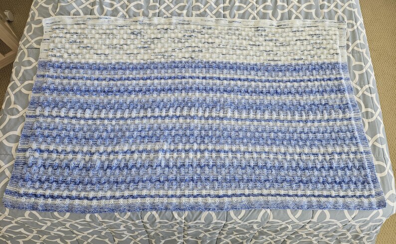 May include: A rectangular, knitted blanket in shades of white and blue. The blanket features a textured pattern with horizontal stripes of varying blue hues, transitioning from lighter to darker shades. The blanket is displayed on a patterned bedspread.