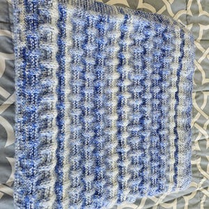 May include: A folded, knitted blanket with a textured pattern in shades of blue, white, and gray. The blanket has a ribbed border and a repeating square design. The soft, cozy blanket is folded on a patterned surface.