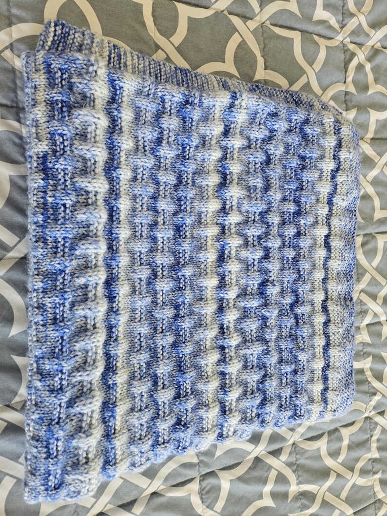 May include: A folded, knitted blanket with a textured pattern in shades of blue, white, and gray. The blanket has a ribbed border and a repeating square design. The soft, cozy blanket is folded on a patterned surface.
