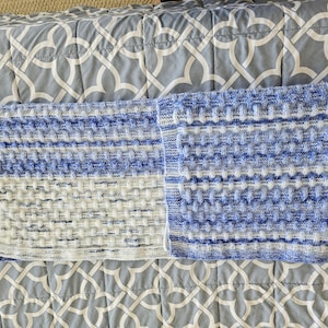 May include: A blue and white knitted blanket with a textured pattern. The blanket is folded in half, revealing a detailed stitch design. The color scheme includes shades of blue and white, creating a soft, inviting aesthetic. The blanket is placed on a patterned gray surface.