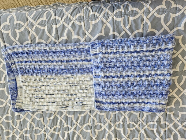 May include: A blue and white knitted blanket with a textured pattern. The blanket is folded in half, revealing a detailed stitch design. The color scheme includes shades of blue and white, creating a soft, inviting aesthetic. The blanket is placed on a patterned gray surface.