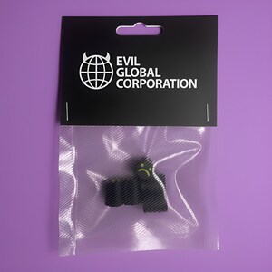 Puede incluir: Un paquete negro con el texto "EVIL GLOBAL CORPORATION" y un logotipo de un globo con cuernos. Dentro de la bolsa de plástico transparente hay una figura negra con una cara triste amarilla. El fondo es de color morado liso.