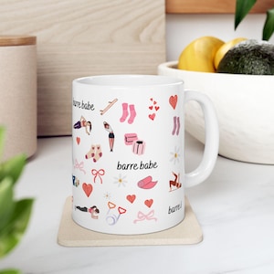 Könnte beinhalten: Weiße Keramiktasse mit einem sich wiederholenden Muster von Ballettthemen-Illustrationen, darunter Tänzer, Socken, Herzen, Schleifen und der Text "barre babe". Die Tasse steht auf einem hellen Untersetzer.