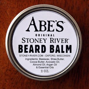 May include: A silver tin with a white label that reads "Abe's Original Stoney River™ Beard Balm". The label also includes the website "stoney-river.com", the location "Oxford, Wisconsin", and a list of ingredients: beeswax, shea butter, cocoa butter, avocado oil, almond oil, argan oil, and essential oils. The tin is 2 oz.