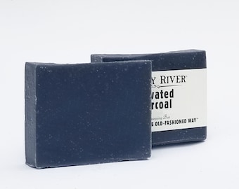 May include: Two bars of dark gray activated charcoal soap. The bar on the right has a white label that reads "Clay River" and "Activated Charcoal Cleansing Bar The Old-Fashioned Way."