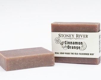 May include: Two bars of Stoney River Cinnamon Orange real soap made the old-fashioned way. The soap is a light brown color and has a rough texture. The bar on the right has a white label with the brand name and product name.