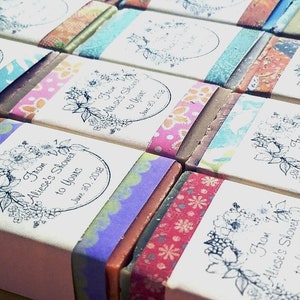 May include: Close-up of several wrapped soaps. Each soap is wrapped with a patterned band and a label with the text "From Aliese's Shower to Yours June 30, 2018" and a floral design. The bands feature various colors and patterns.