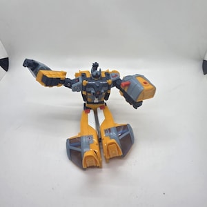 May include: A yellow, gray, and black toy robot in a dynamic pose. The robot has articulated arms and legs, with a detailed design featuring panels and mechanical elements. The toy is set against a plain white background.