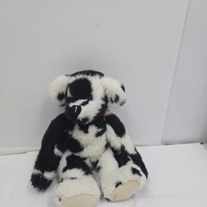 May include: A plush teddy bear featuring a black and white cow-print pattern across its soft, fuzzy fur. The bear has rounded ears, a dark stitched nose, and light-colored fabric on the soles of its feet, with a small red accent visible at the neck.