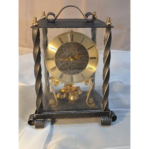 May include: A vintage clock with a gold-tone face and black Roman numerals. The clock is encased in a clear glass box with a black wrought iron frame and a decorative handle. The clock's pendulum is gold-toned.