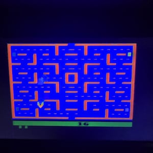 May include: A screenshot of the classic arcade game Pac-Man. The game features a blue maze with orange pathways and a green base. The number "16" is visible at the bottom. The Pac-Man character and ghosts are also visible.
