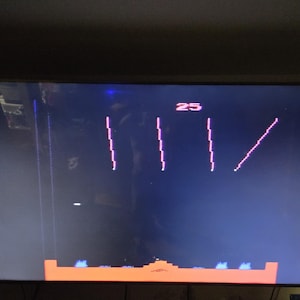 May include: A screenshot of a retro video game on a dark screen. The game features vertical lines and a diagonal line in pink, with the number 25 at the top. A horizontal orange bar with blue accents is at the bottom.