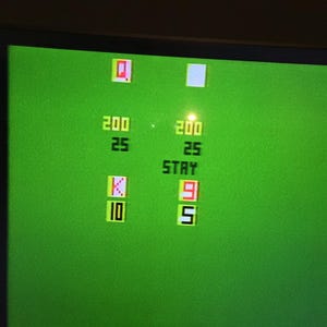 May include: A green screen displays a retro video game interface. The screen shows the numbers 200 and 25, a red Q, a white square, the word STAY, a red 9, a 5, a K, and a 10. The game has a simple, pixelated design.