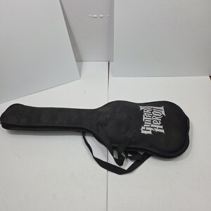 May include: A black soft-sided guitar case featuring the white text Guitar Hero printed on the front. The case includes a long neck section, a wider body section, and black fabric carrying straps attached to the exterior of the main compartment.