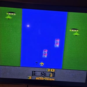 May include: A screenshot of a retro video game with a blue river separating two green land masses. A yellow plane flies over the water, with two buildings on each side. The game displays the word "FUEL" and the word "ACTIVISION".
