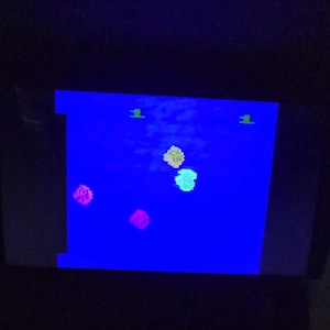 May include: A screen showing a retro video game with a blue backdrop. The game features colourful pixelated shapes and the number "1" in green at the top. The shapes are pink, yellow, and light blue.