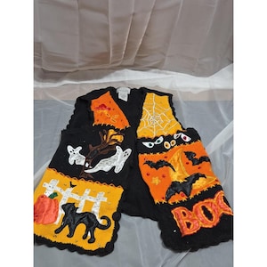May include: Black Halloween vest with orange and yellow accents. Features a pumpkin, black cat, ghosts, bats, and the word "BOO". The vest is decorated with a spider web, stars, and a witch's hat.