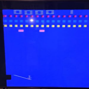 May include: A screenshot of a classic video game with a blue background. The game features rows of colored blocks in red, yellow, and blue, with a paddle at the bottom and a ball. The game is a retro arcade game.