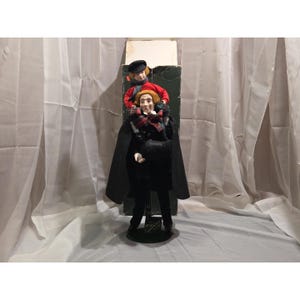 May include: A decorative doll featuring two figures. One figure is wearing a black coat and pants, and the other is wearing a red shirt and black hat. Both dolls have red hair and are standing in front of a green box.