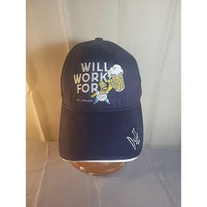 May include: Navy blue baseball cap featuring the phrase "WILL WORK FOR" in white lettering, along with an image of Homer Simpson holding a beer. The cap has a white trim and a signature on the side.