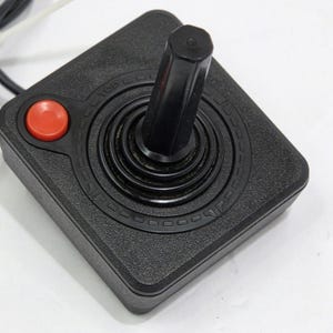 May include: A black video game joystick controller with a square base, a single red circular button, and a central vertical stick. The base features a textured surface with the word TOP embossed near the button and a black cable extending outwards.