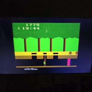 May include: A screenshot of an old video game with a green background, brown trees, and a yellow platform. The game displays the score 1775 and the time 19:44. The Activision logo is at the bottom.