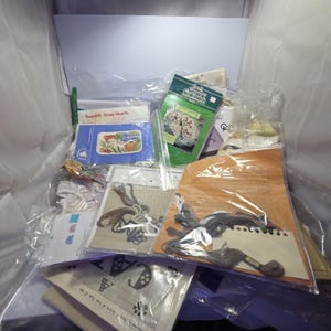 May include: A selection of craft kits and supplies, including cross-stitch patterns, embroidery threads, and packaged needlework projects. The kits feature floral and heart designs, with various colours and textures visible. The items are in clear plastic packaging.