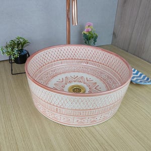 Pode incluir: Uma pia redonda de cerâmica com um padrão detalhado em rosa e branco. A pia tem um ralo dourado e está sobre uma superfície de madeira clara. Uma torneira de cobre está no fundo. O diâmetro da pia é de cerca de 40 cm.