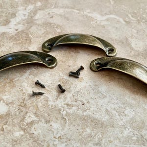 May include: Three antique brass cabinet pulls with a curved shape and a smooth finish. The pulls are accompanied by five small black screws.