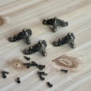 May include: Four antique bronze-colored furniture feet with ornate, curved designs and a central teardrop shape. Small black screws are scattered around the feet on a light wood surface. These decorative feet are ideal for furniture restoration or crafting projects.